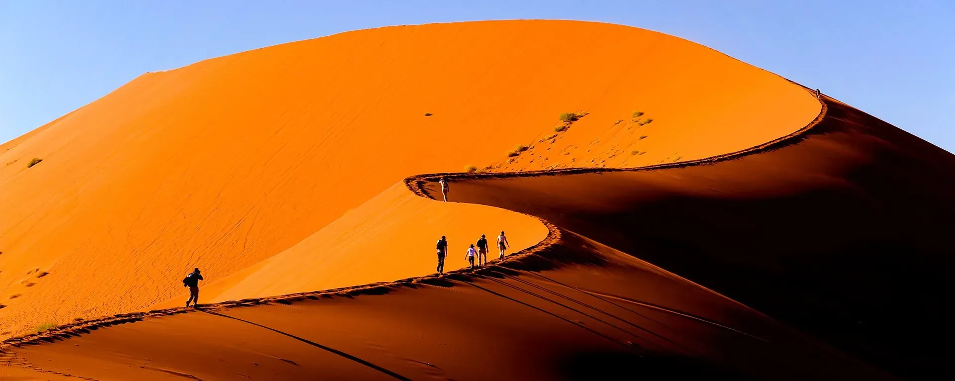 people trekking across towering orange sand dunes under a clear blue sky during a dune adventure