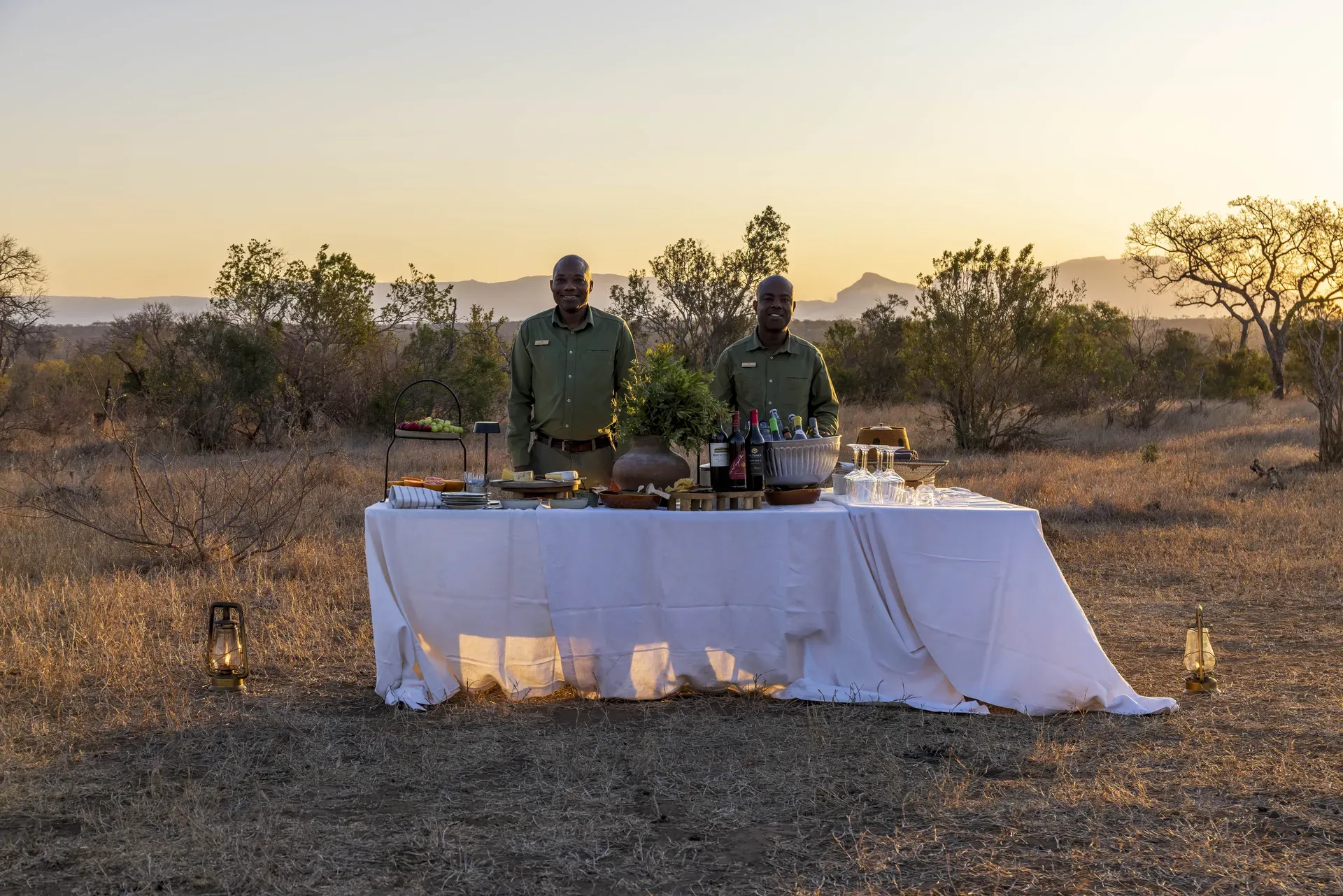 Table set with food and wine in the African bush at Monwana Lodge, with two guides standing behind it.