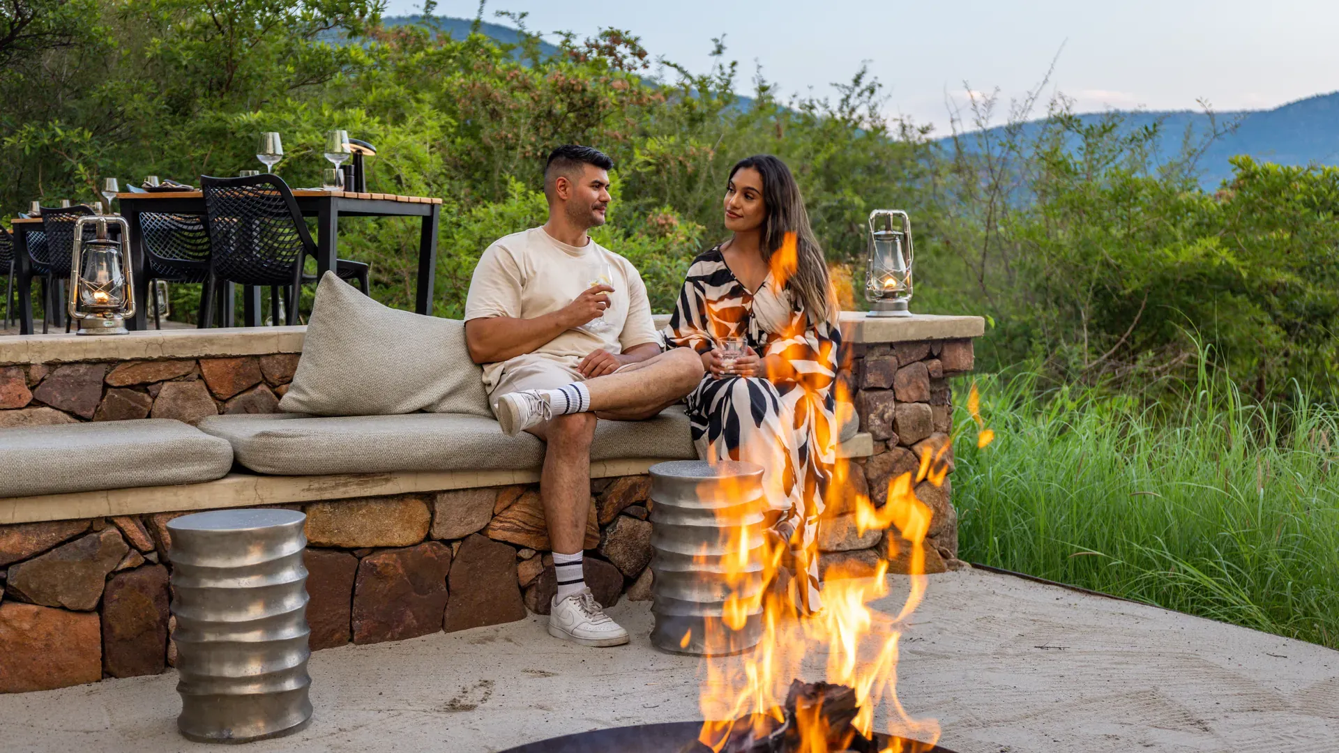Couple enjoying an intimate outdoor dining experience around a fire pit with a small table and drinks in a lush garden