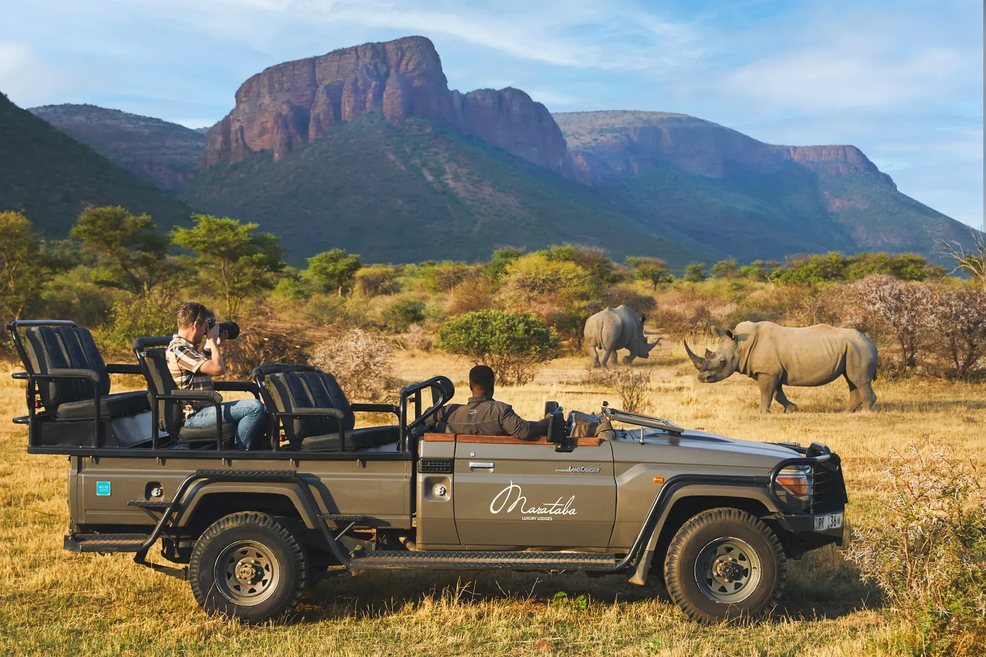 Safari vehicle driving through open savanna at the foot of a mountain under a clear blue sky